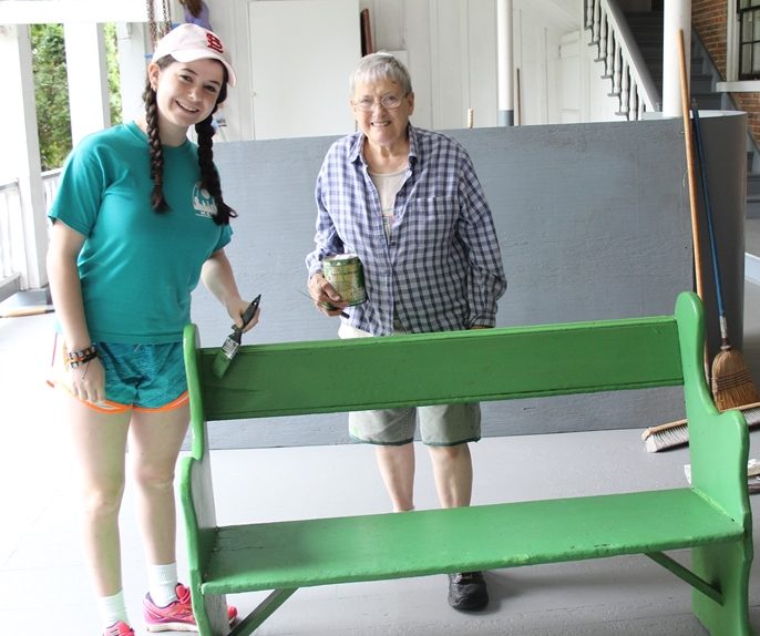A young woman and a Sister hold a paint brush and can of paint as they stand next to a green bench.