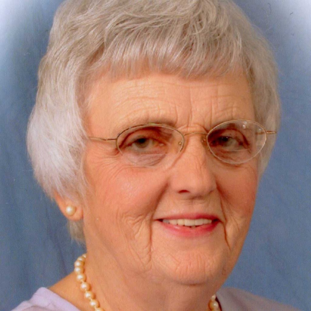 A woman with short grey hair and glasses wearing pearl earrings, a pearl necklace, and a lilac blouse smiling for a headshot picture with a blue and white vignette background.