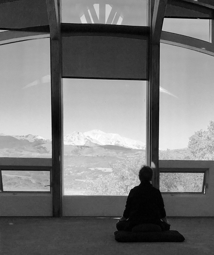 A person meditates in front of large windows with a mountain view in the background