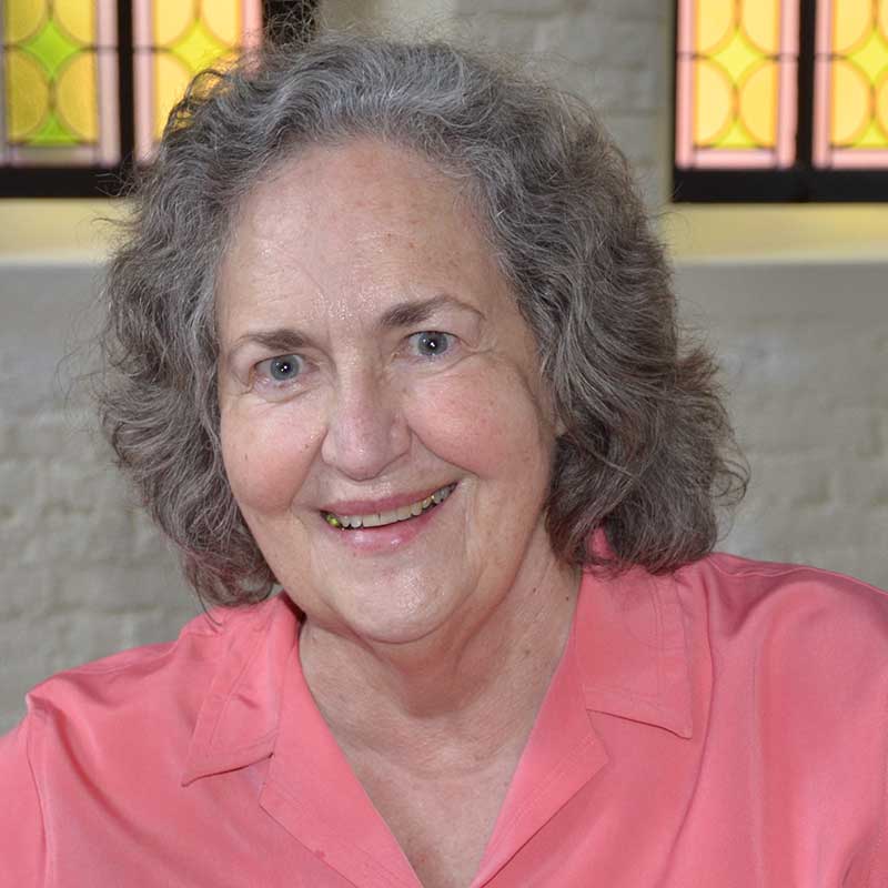 A woman with shoulder length brown and grey hair and blue eyes wearing a pink blouse smiling for a headshot picture indoors with a white brick and stained glass window background.