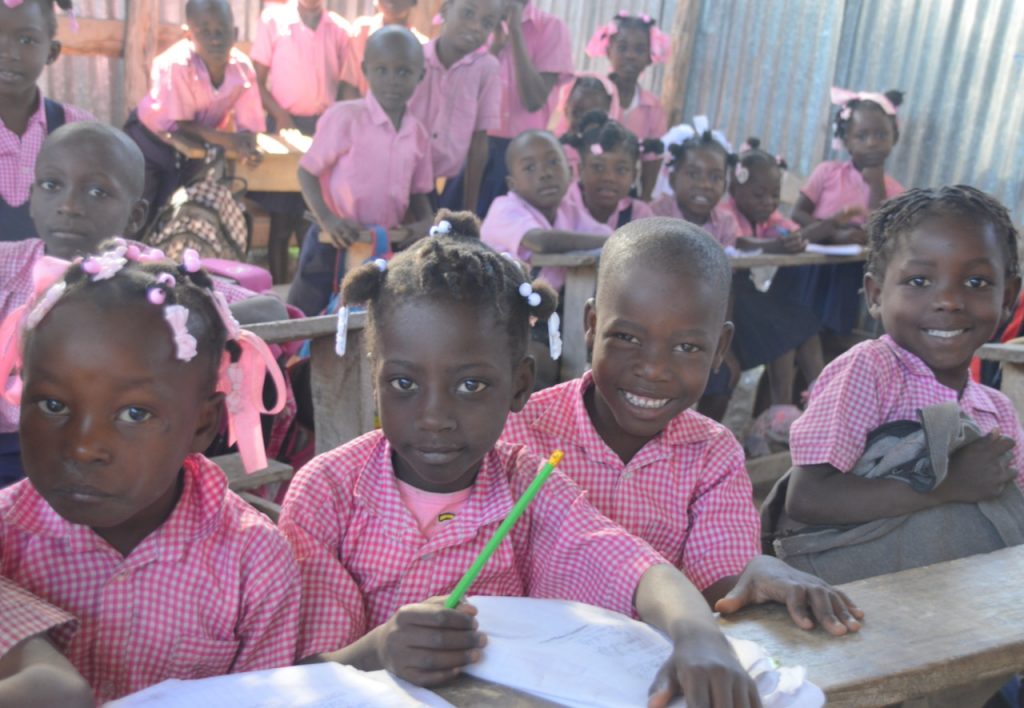 Haitian students in school uniforms sit at a shared desk in their outdoor classroom.