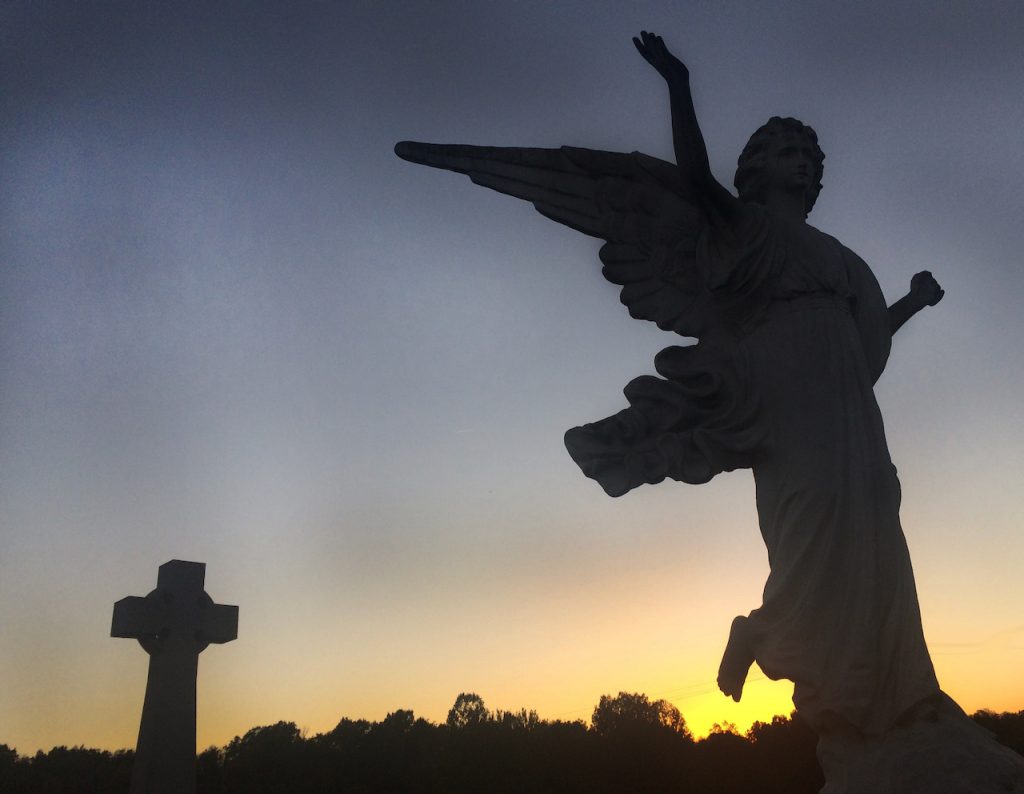 An angel and a cross are profiled in front of the setting sun.