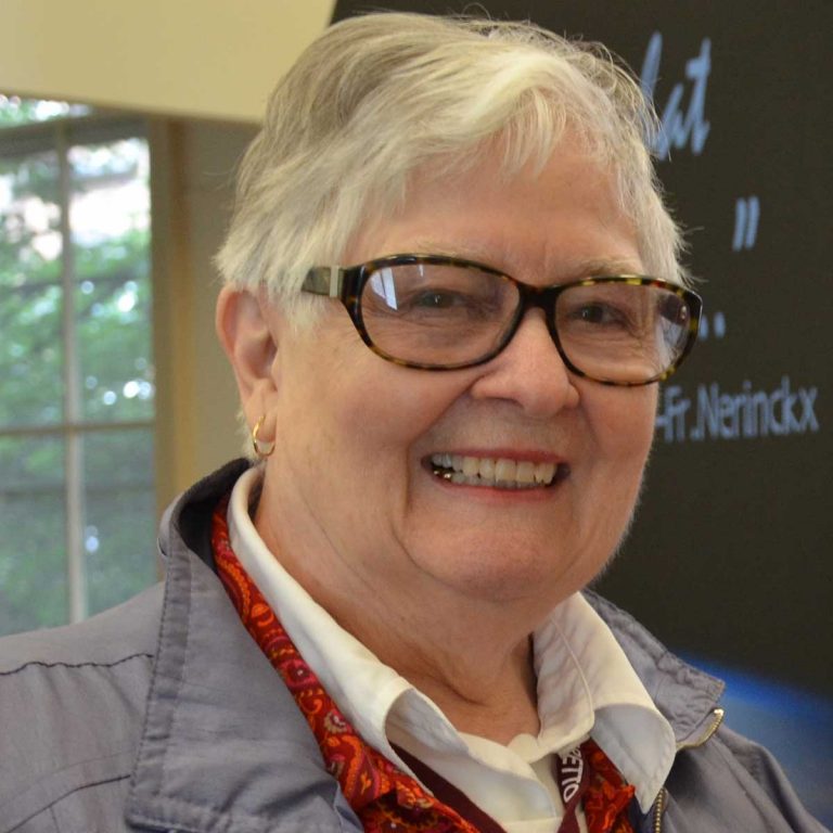 A woman with short grey hair and black-framed glasses wearing a white collared blouse, a red sweater and a grey jacket smiling brightly for a headshot picture indoors.