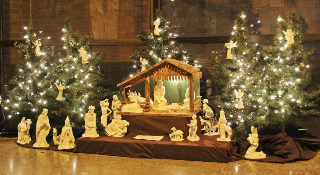 A manger scene is on display, surrounded by small evergreens sparkling with white lights