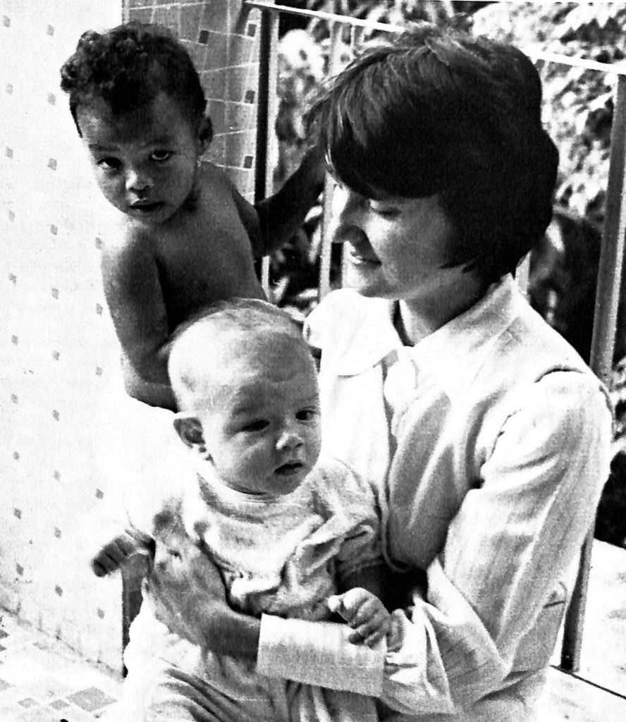 In this black-and-white archival photo, a woman holds an infant as a toddler stands next to her.