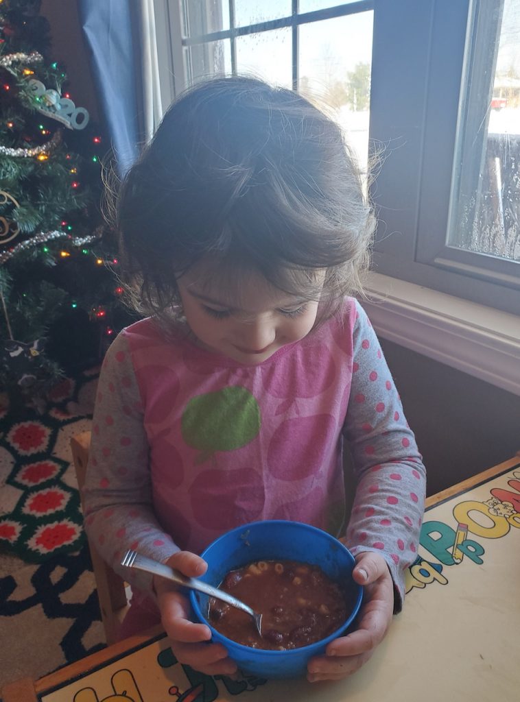 A little girl sits with a bowl of chili. A Christmas trees is behind her and snow can be seen through the windows.