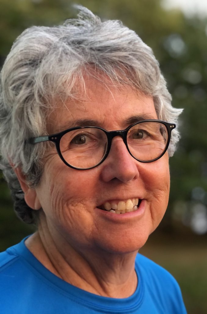 A woman, Eileen Harrington with short grey hair and dark-framed glasses smiling brightly for a headshot picture outdoors wearing a blue t-shirt.