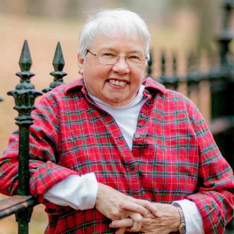 A woman with short white hair and wire-framed glasses wearing a long sleeve white undershirt and a red and green plaid collared button-up shirt smiling for a portrait picture while posing by resting her elbow on an iron fence.