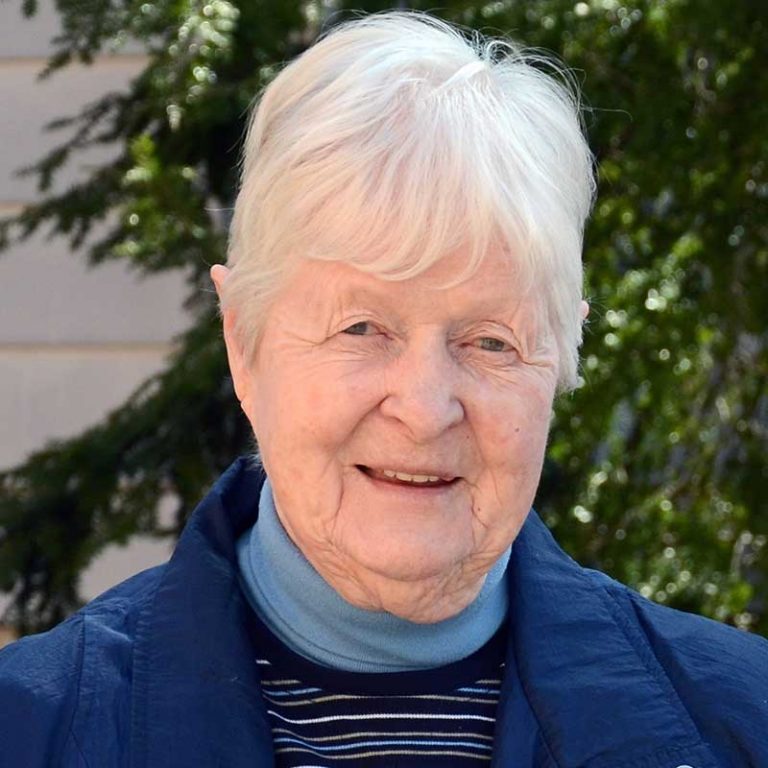 A woman with short white hair wearing a blue turtleneck, a dark-colored striped sweater, and a blue jacket smiling brightly for a picture outdoors.