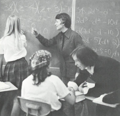 Black-and-white archival photo: A woman instructs a student at a blackboard doing math equations while two other students confer in the foreground.