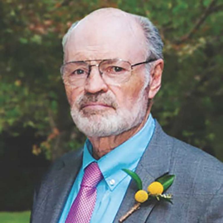 A man with short grey hair, a grey beard and wire-framed glasses smiling softly for a portrait picture outdoors wearing a blue dress shirt, purple tie, and a dark grey suit jacket with a yellow berry pin on the chest.