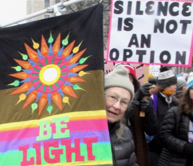 Woman in a parade carries a colorful banner that reads "Be Light..." Photo by Philip Deitch