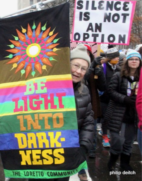 Woman in a parade carries a colorful banner that reads "Be Light Into Darkness." Photo by Philip Deitch