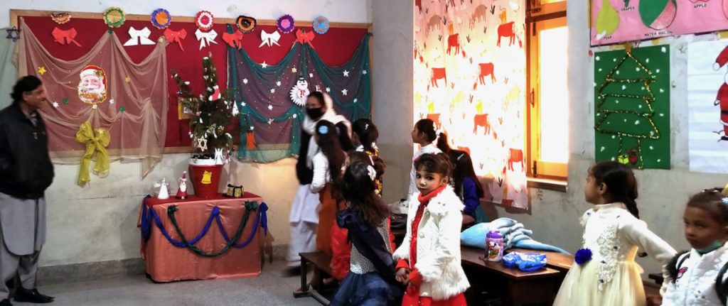 An elementary classroom in Pakistan displays a Christmas tree and other Christmas decorations. A man and woman in their shalwar kameez stand at the front of the room.