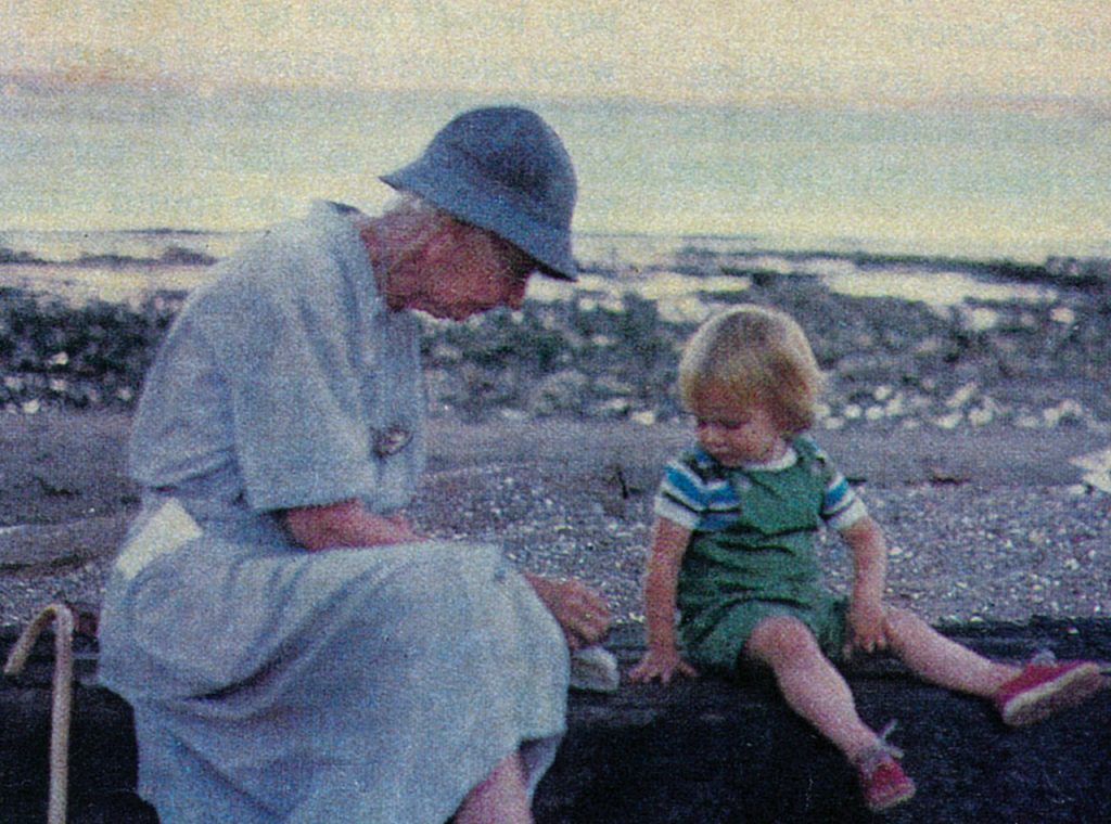 An older woman, with a cane propped behind her, stacks pebbles on the log on which she sits, while the toddler next to her watches.