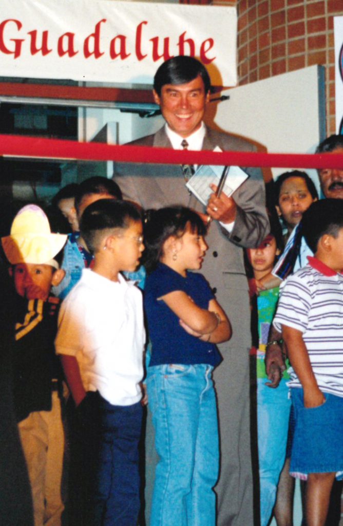A man stands with a crowd of children behind a red ribbon as he prepares to cut it.