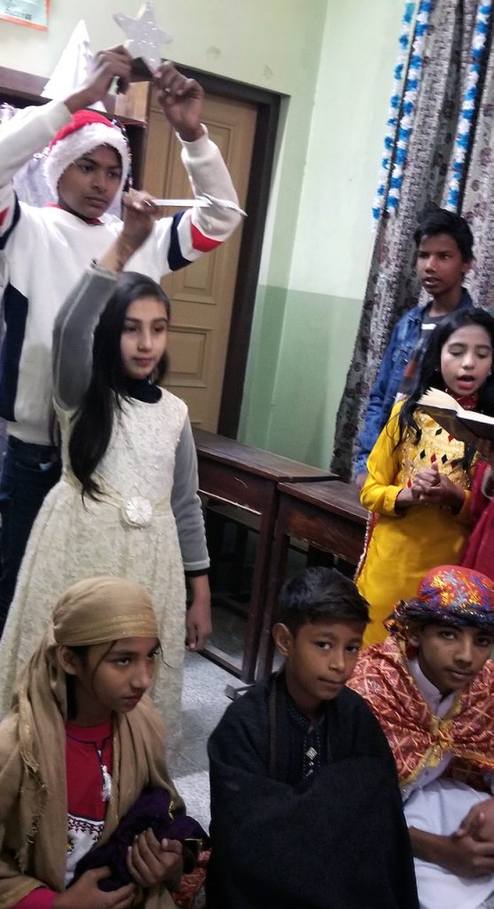 Pakistani elementary students act out the Nativity story, with one holding a star, one portraying an angel, two as Mary and Joseph, and several others in colorful costumes.