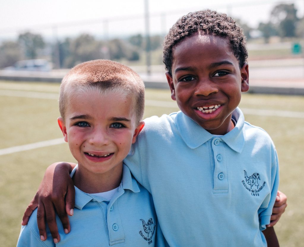 Two boys, one white, one Black, wearing school uniforms, stand with arms around each other's shoulders.