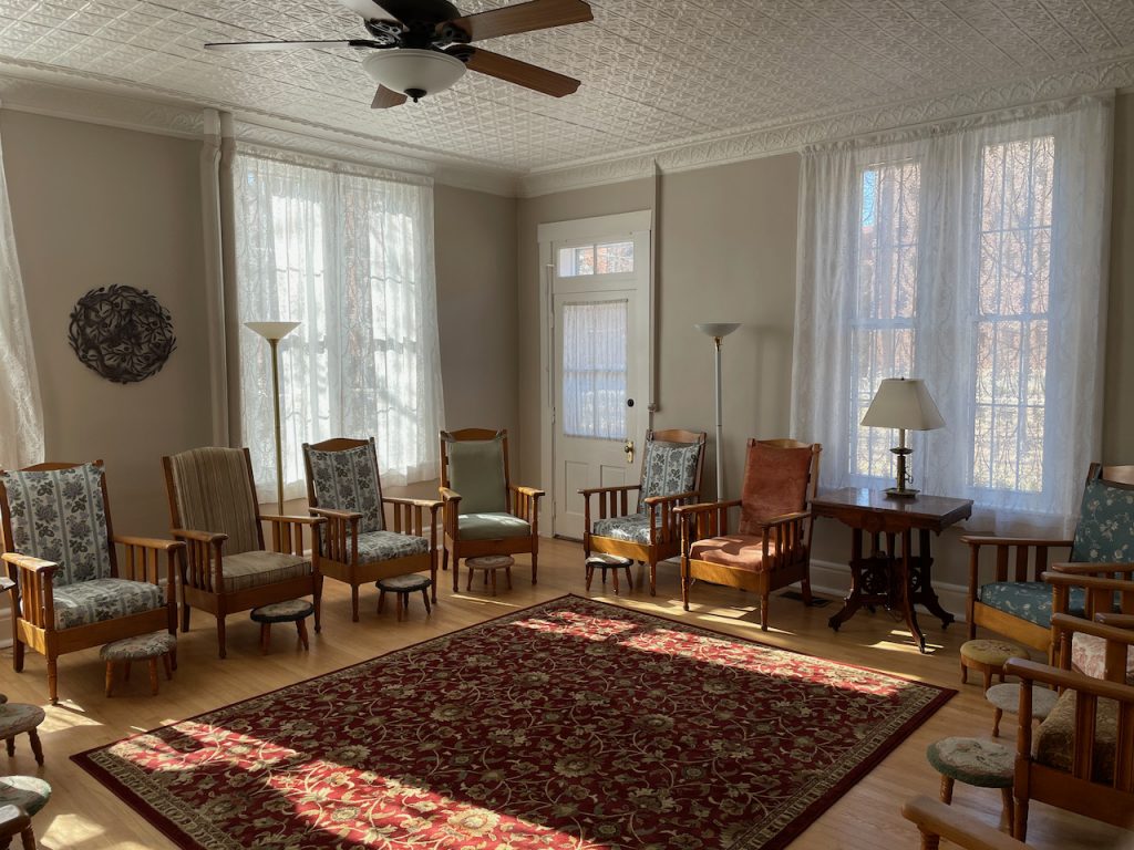 Comfortable chairs are arranged in a circle around the edges of a large rug in a bright and airy room with a punched-tin ceiling.