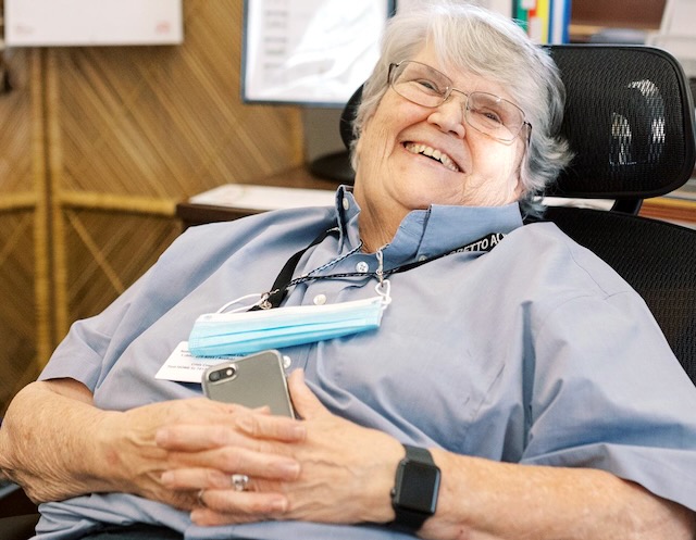 Buffy Boesen SL sits back in a chair with hands clasped holding a phone. She's wearing a blue shirt and glasses.