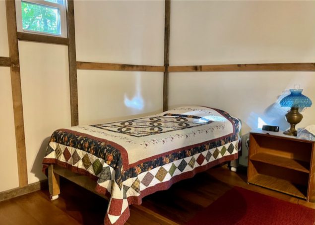 A twin bed with a quilt is positioned in the corner of the room. A small window is up to the left, and a bedside table with lamp is on the right.
