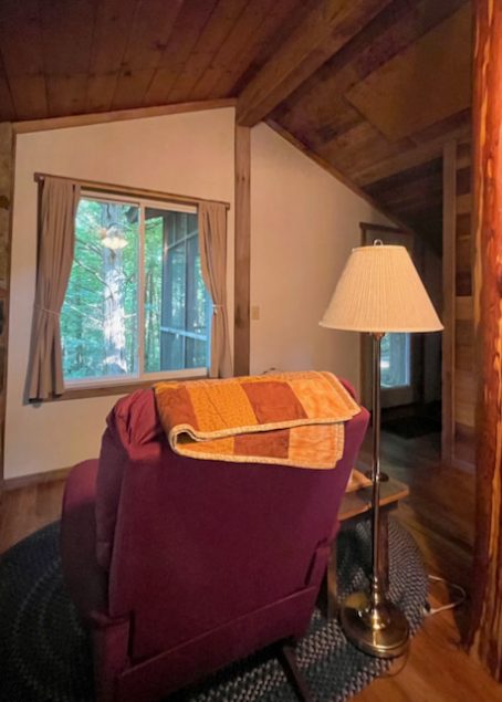 An easy chair faces out a window with a view of the forest. A floor lamp stands next to the chair.