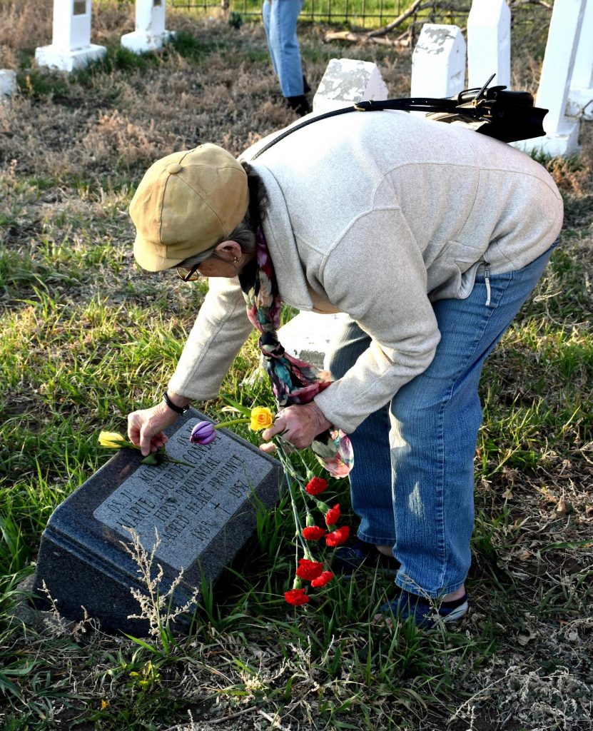A woman wearing a brimmed hat places a single flower on a grave marker.