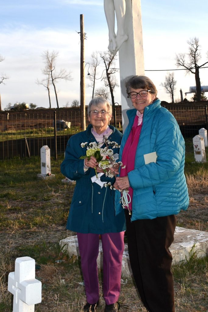 Two women holding flowers stand at the foot of a large crucifix. A small stone cross is in the foreground and additional simple headstones are visible in the background.