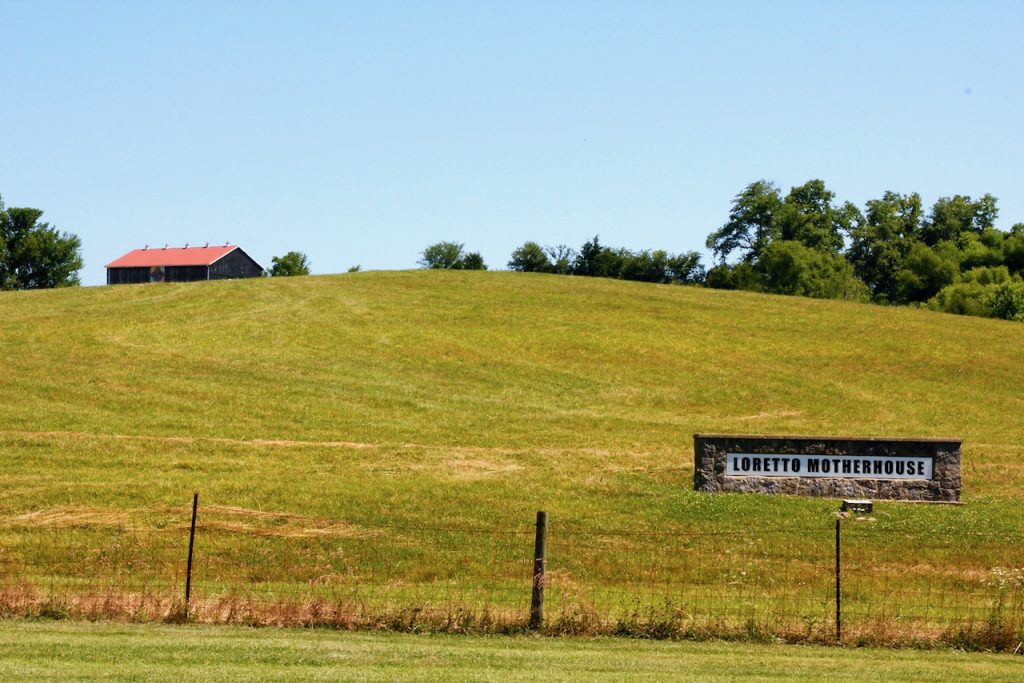 An entrance sign reading "Loretto Motherhouse" sits at the bottom of a grassy hill. The roof of a building is visible at the top of the hill.