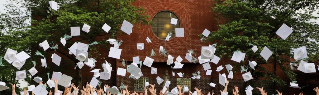 Hands toss white graduation caps with green tassels into the air.