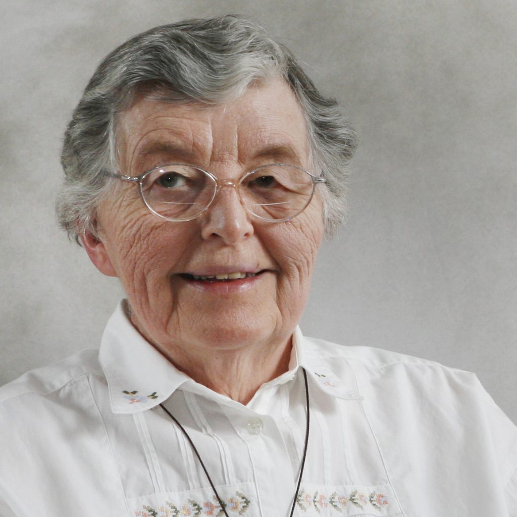A woman with short dark grey hair and round glasses wearing a white collared blouse and long pendant necklace smiling for a headshot portrait picture with a plain light grey background.