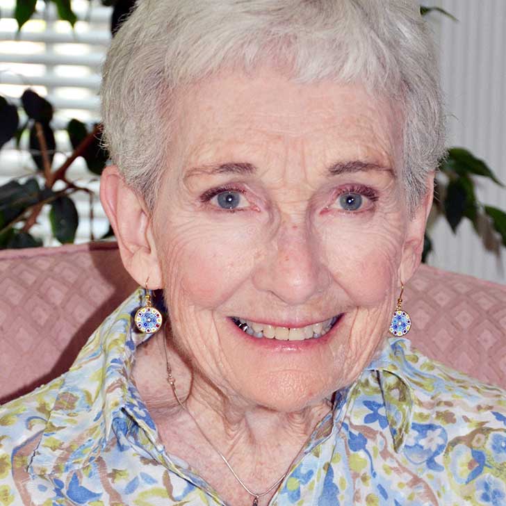 A woman with short grey hair and dangly earrings wearing a light-colored patterned blouse smiling brightly for a headshot picture while sitting in a plush pink chair in a living room with a house plant branch behind her head.