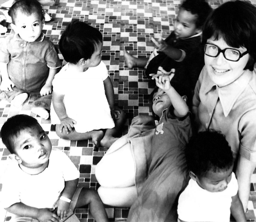 Black-and-white photo of a White woman sitting on the floor with six toddlers of varying racial backgrounds playing around her.