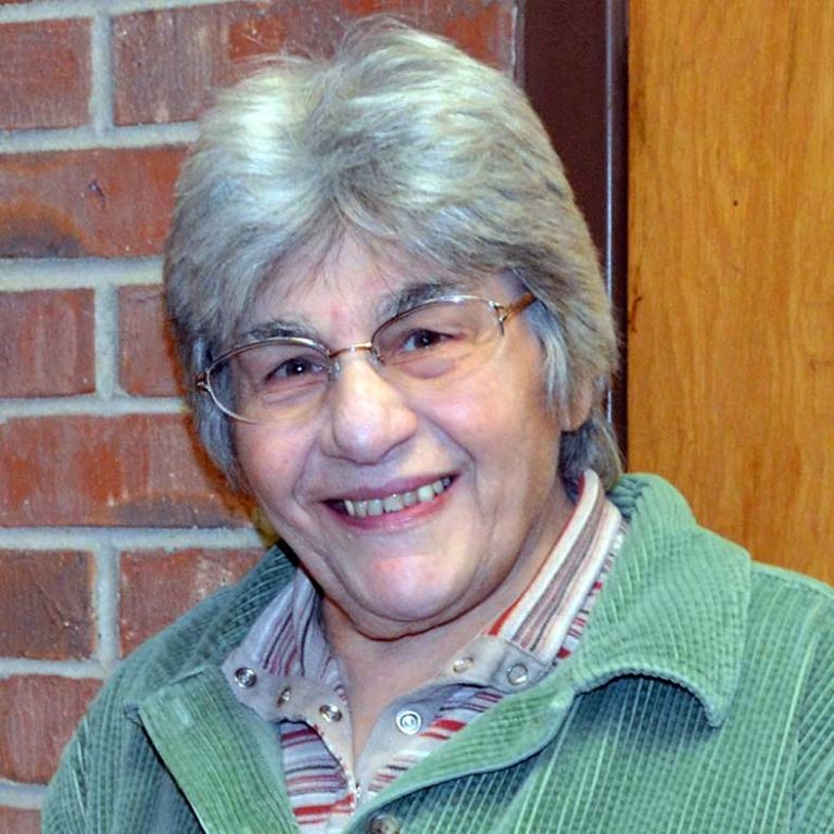 A woman with short grey hair and glasses wearing a striped collared shirt and a bright green jacket smiling for a headshot picture next to a brick building.