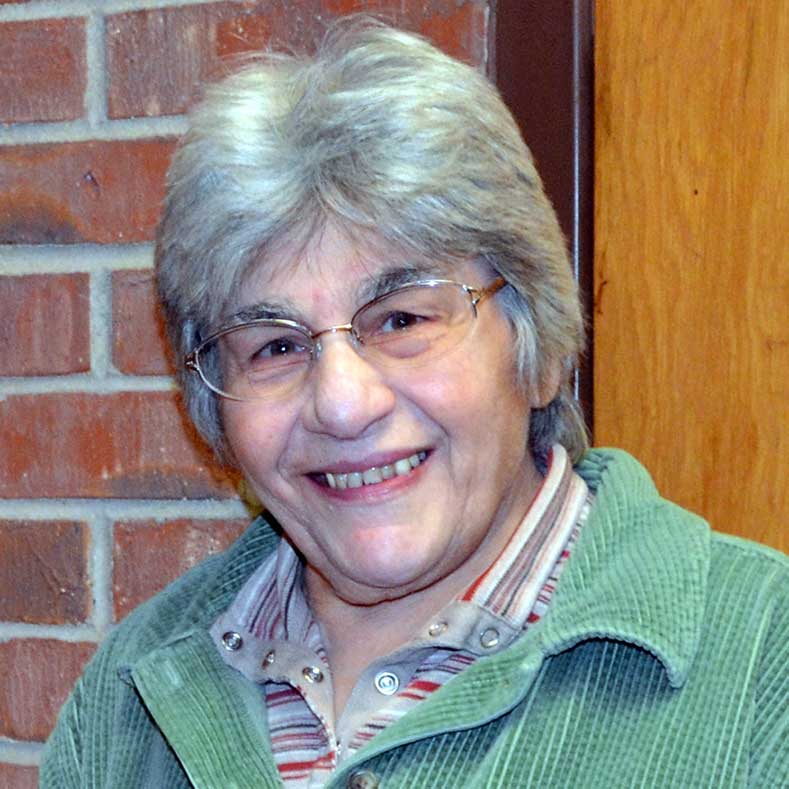 A woman with short grey hair and glasses wearing a striped collared shirt and a bright green jacket smiling for a headshot picture next to a brick building.