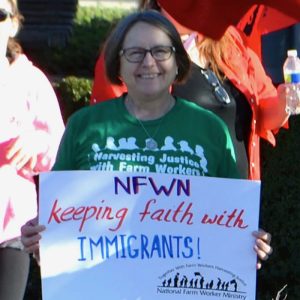 A woman wearing a green shirt and a smile stands amongst a crowd and holds a sign that reads "NFWN keeping faith with Immigrants", with the National Farm Worker Ministry logo in the lower right corner.