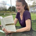 Jessie Rathburn reads "Braiding Sweetgrass" at an outdoor table, with gardens and a shed in the background.