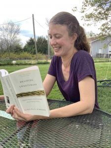 Jessie Rathburn reads "Braiding Sweetgrass" at an outdoor table, with gardens and a shed in the background.