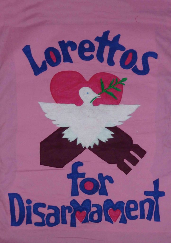 A pink banner with a white dove in the middle has blue lettering around it saying "Lorettos for Disarmament"