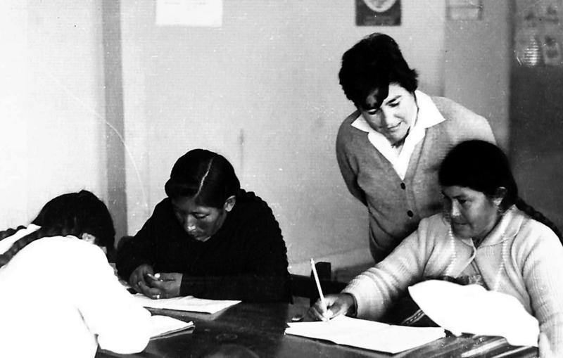 A woman bends over to assist her student with her writing in this archival photo.