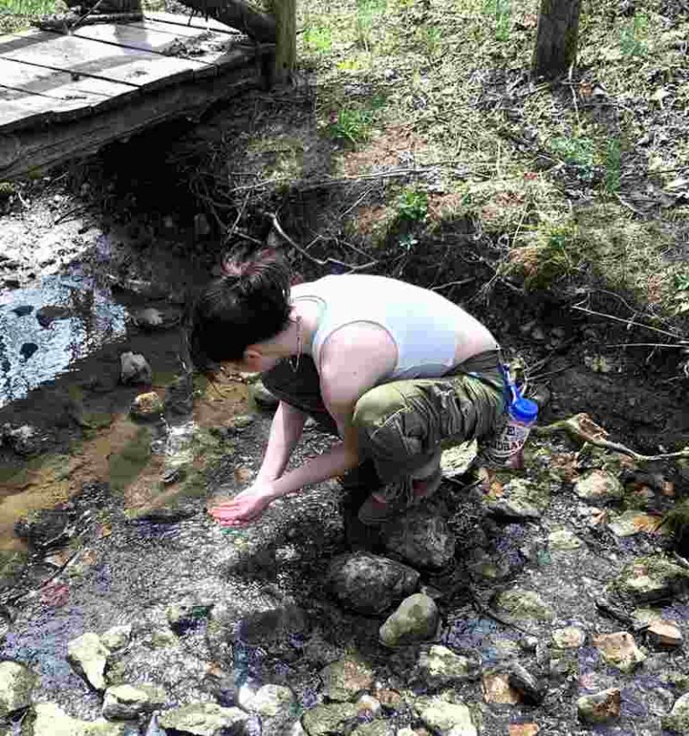A woman squats on a rock in a stream, her hands cupped in front of her.