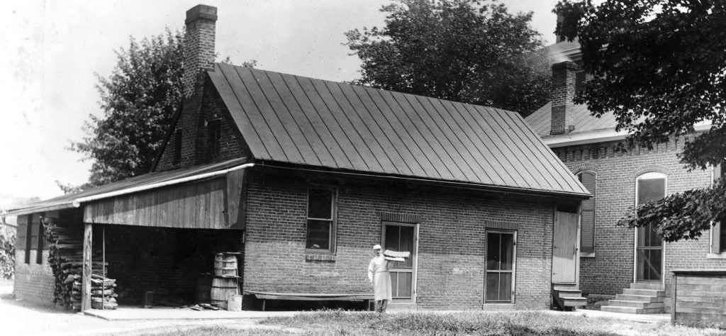 Archival black and white photo of a long brick building with a woodshed attached on the left side. A man with a white hat and long white apron stands in front of the door of the building. 