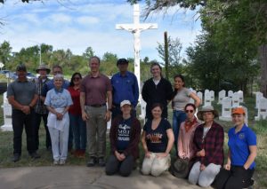 Fourteen men and women pose in front of the Loretto Heights Cemetery, with the large white crucifix and gravestones visible in the background.