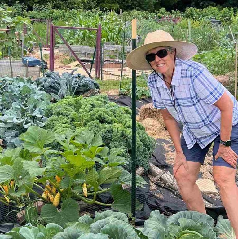A woman in a sunhat, sun glasses and shorts leans in with a smile over a burgeoning garden.