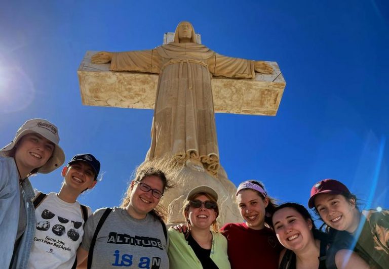 Seven smiling people look down to the camera. A statue of the risen Christ on the cross towers above them in the clear blue sky.