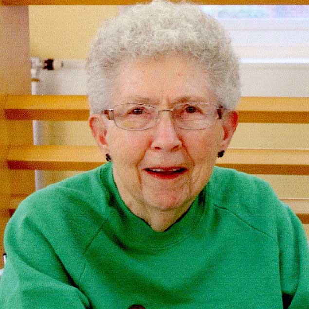A woman with short curly white hair and glasses wearing a bright green sweater and black stud earrings smiling for a headshot picture indoors.