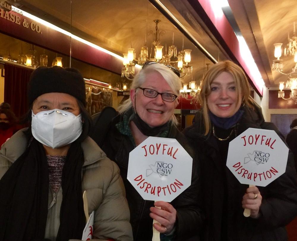 Three women (one masked, the other two holding signs reading "Joyful Disruption") pose together for a photo.