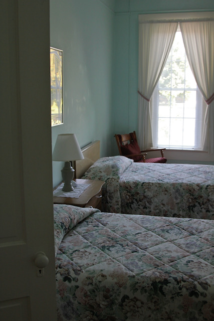 Two twin beds, a table and lamp between them, are neatly made up with floral quilts. Sunlight streams through the window behind them.