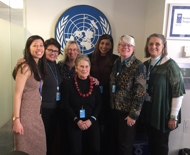 Seven women pose for a photo in a room displaying the United Nations logo.
