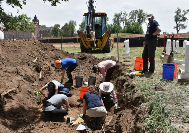 Six people carefully dig in a large space in a cemetery cleared by a backhoe. One woman stands on the side amidst white gravestones and observes.